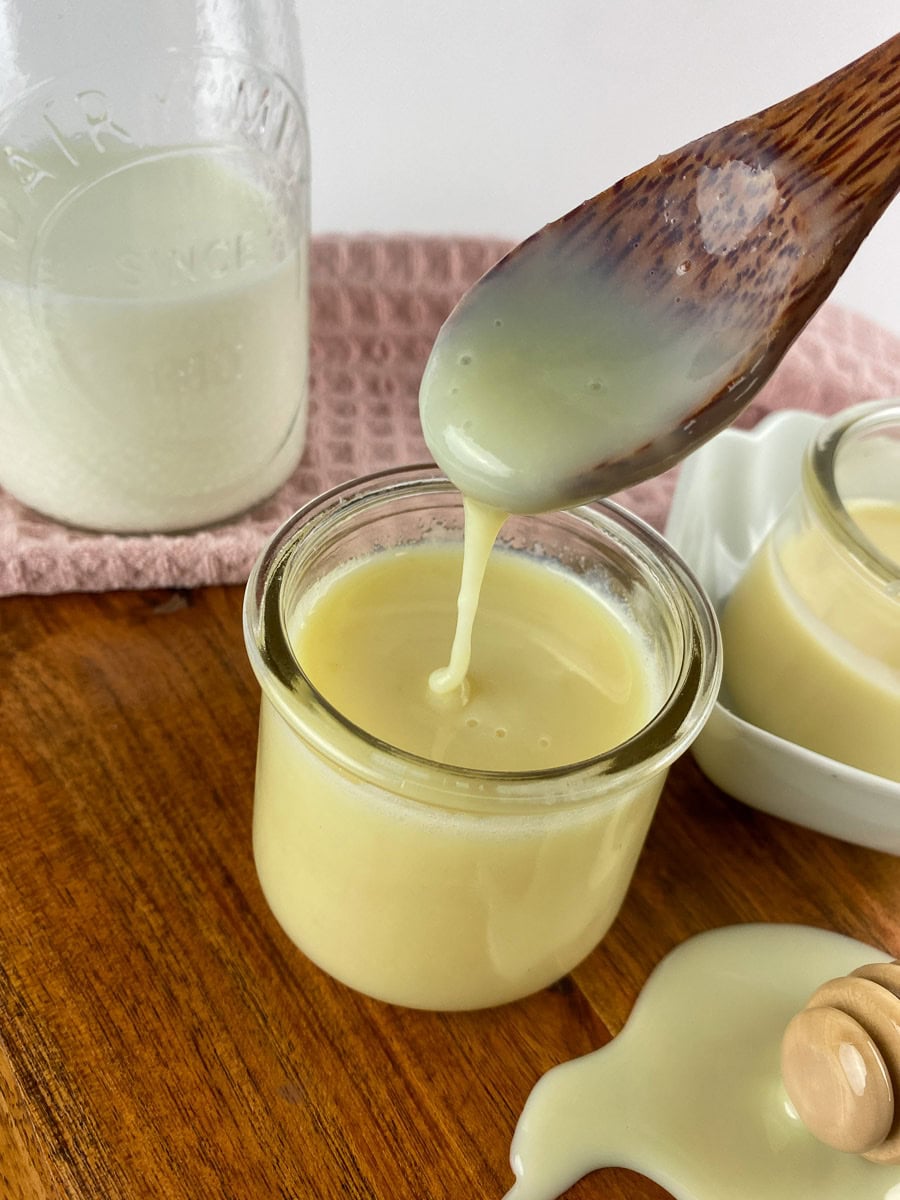 Wooden spoon drizzling homemade sweetened condensed milk into a glass jar on a wooden table, with a pink cloth and milk bottle in the background.