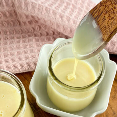 Homemade sweetened condensed milk in a glass jar with a wooden spoon drizzling the creamy milk, placed on a wooden board with a pink cloth in the background.