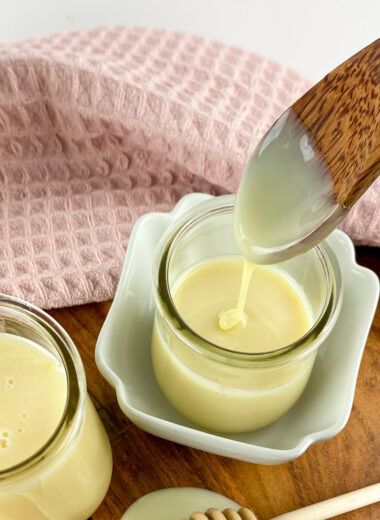 Homemade sweetened condensed milk in a glass jar with a wooden spoon drizzling the creamy milk, placed on a wooden board with a pink cloth in the background.