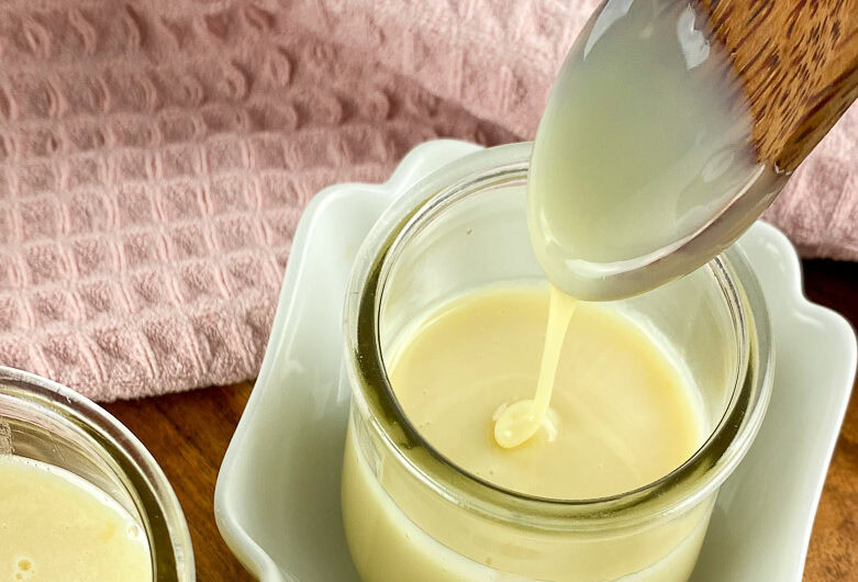 Homemade sweetened condensed milk in a glass jar with a wooden spoon drizzling the creamy milk, placed on a wooden board with a pink cloth in the background.